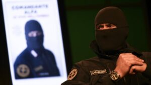 Comandante Alfa in uniforme nera e passamontagna, seduto davanti a un suo ritratto
